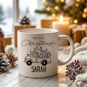 May include: A white ceramic mug with a black graphic of a truck carrying a Christmas tree. The mug has the words "MERRY CHRISTMAS" and the name "SARAH" printed on it. The mug is surrounded by Christmas decorations.