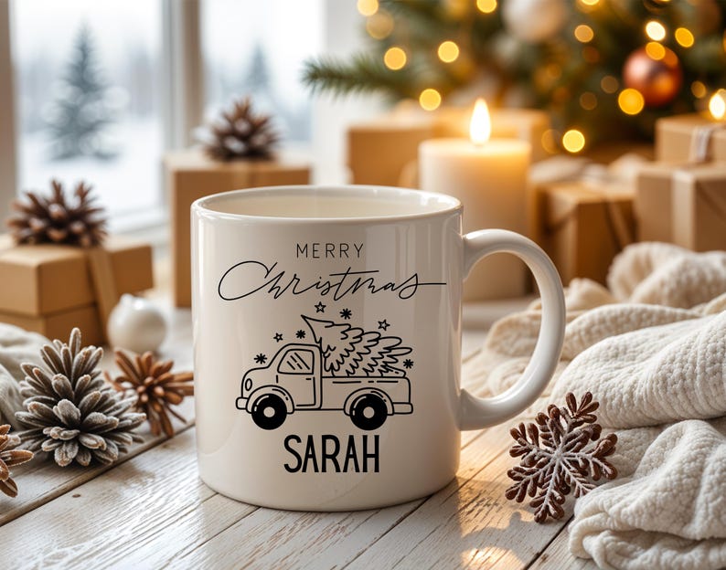 May include: A white ceramic mug with a black graphic of a truck carrying a Christmas tree. The mug has the words "MERRY CHRISTMAS" and the name "SARAH" printed on it. The mug is surrounded by Christmas decorations.