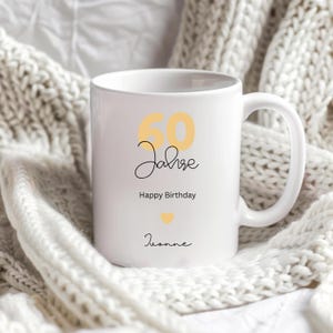 May include: White ceramic mug with the text "60 Jahre Happy Birthday Joanne" in black and yellow script. A yellow heart is also featured on the mug. The mug is sitting on a white knitted blanket.