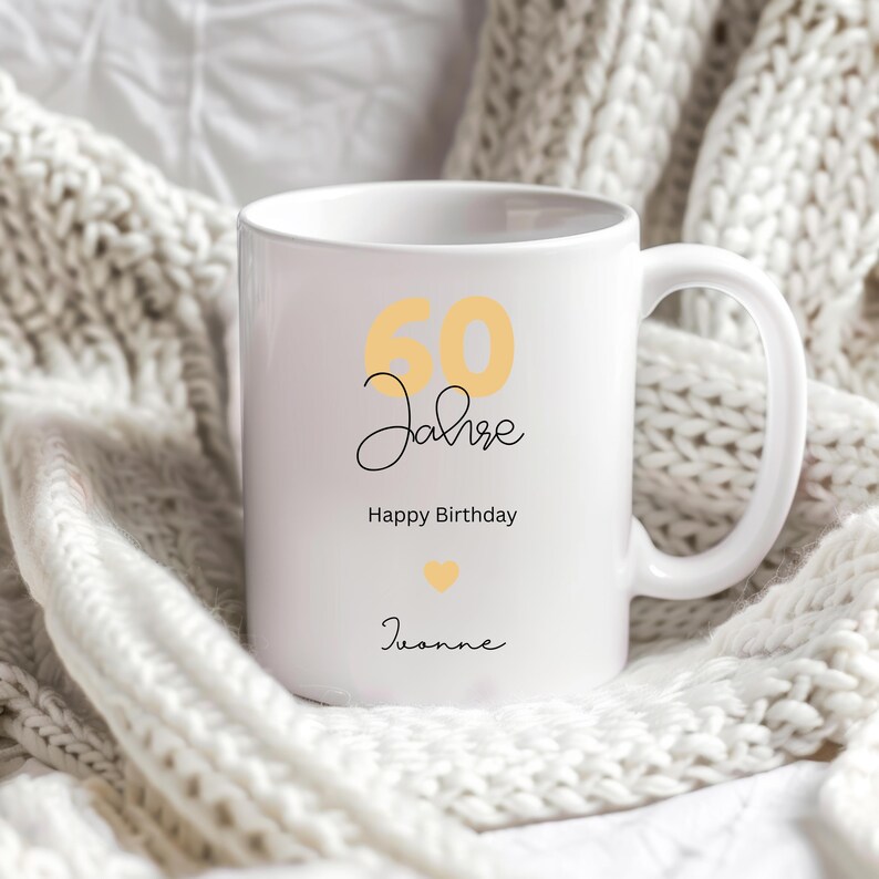 May include: White ceramic mug with the text "60 Jahre Happy Birthday Joanne" in black and yellow script. A yellow heart is also featured on the mug. The mug is sitting on a white knitted blanket.