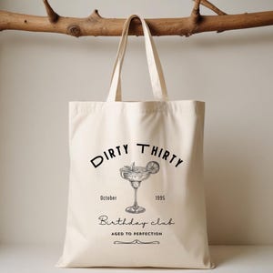 May include: Cream-colored tote bag with black text and a cocktail illustration. The text reads "Dirty Thirty" above a margarita glass, with "October 1995" and "Birthday Club Aged to Perfection" below.