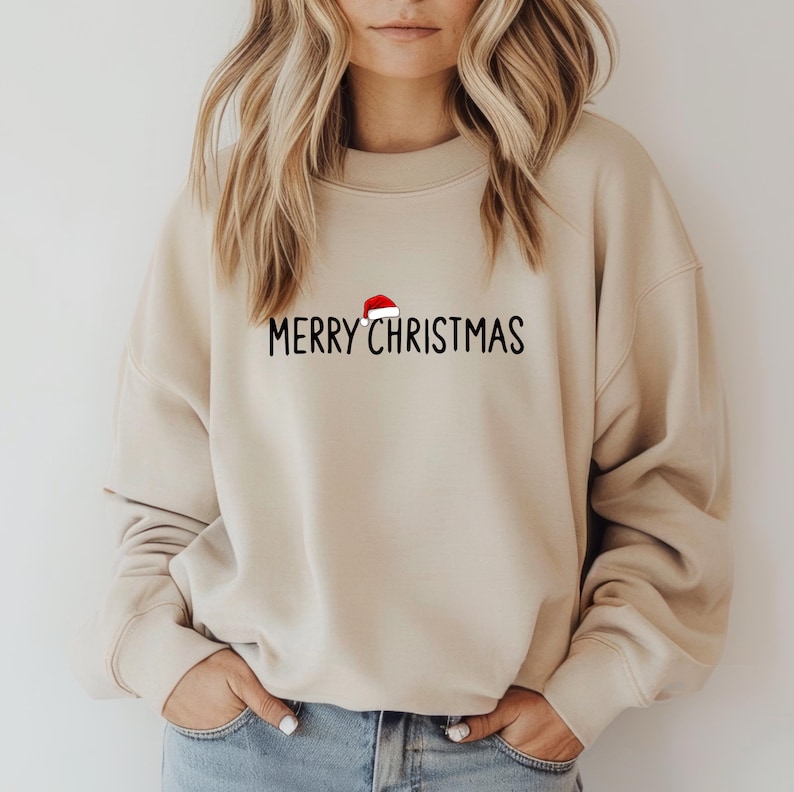 May include: A beige crewneck sweatshirt featuring the black text MERRY CHRISTMAS printed across the chest. A small red and white Santa hat graphic is positioned directly above the letter R in the word MERRY.
