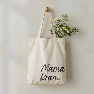 May include: Cream-colored tote bag with the words "Mama Kram." in black script. The bag hangs from a gold hook on a white wall. Fresh flowers with white and yellow blooms are inside the bag.