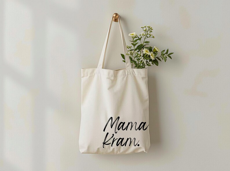 May include: Cream-colored tote bag with the words "Mama Kram." in black script. The bag hangs from a gold hook on a white wall. Fresh flowers with white and yellow blooms are inside the bag.