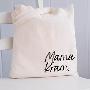 May include: Cream-colored tote bag with the words "Mama Kram" in black script. The bag has two handles and is sitting on a white chair. The bag is made of a soft, natural fabric.