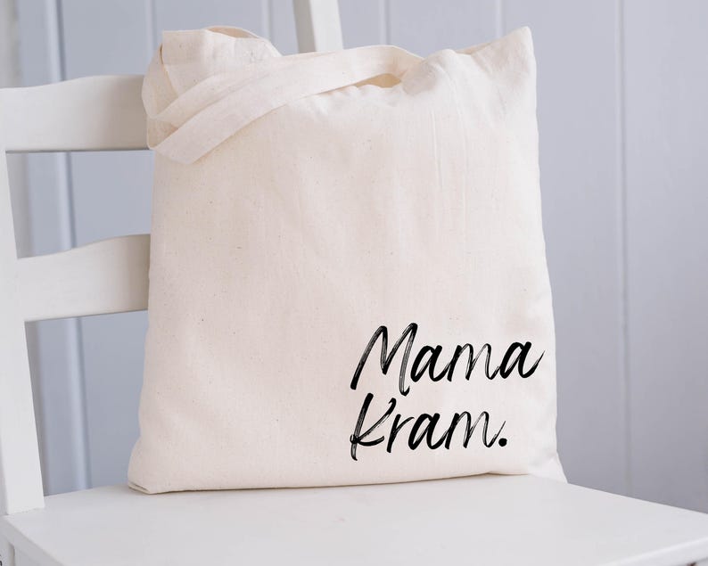 May include: Cream-colored tote bag with the words "Mama Kram" in black script. The bag has two handles and is sitting on a white chair. The bag is made of a soft, natural fabric.