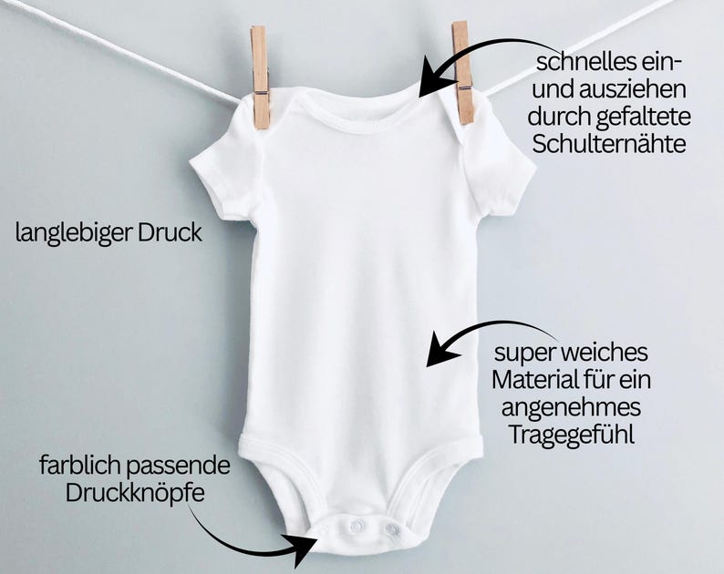 May include: A white baby onesie with short sleeves, hanging from a clothesline with a wooden clothespin. The onesie has snap closures at the bottom. Text on the image says "durable print" and "color-matched snaps".