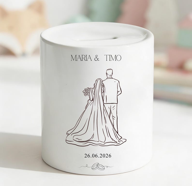 Puede incluir: Hucha de cer&aacute;mica blanca con una ranura en la parte superior. La hucha presenta un dibujo lineal de una novia y un novio, con los nombres "MARIA & TIMO" y la fecha "26.06.2026" impresos en la parte delantera. Dos anillos de boda est&aacute;n dibujados en la parte inferior.