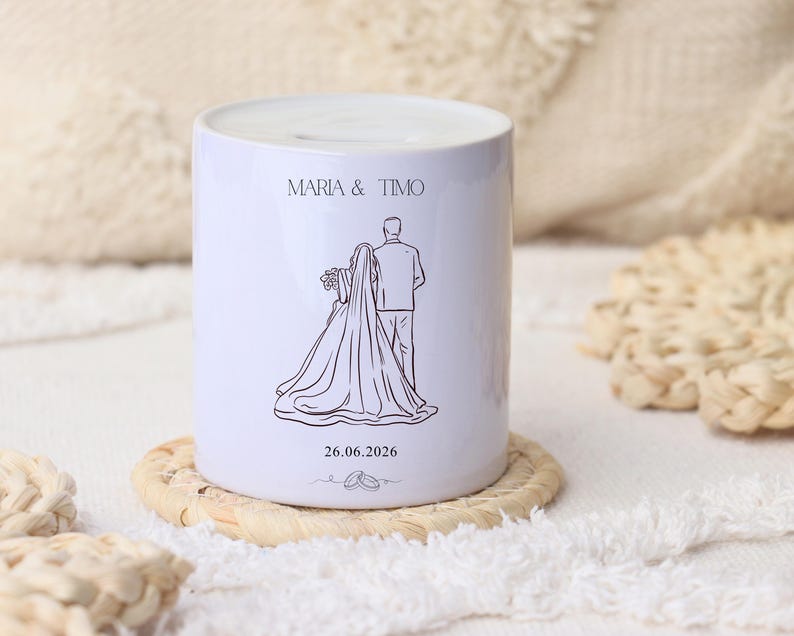 Puede incluir: Hucha de cer&aacute;mica blanca con un dibujo lineal de una novia y un novio, los nombres "MARIA & TIMO" y la fecha "26.06.2026". La hucha est&aacute; sobre un posavasos tejido.