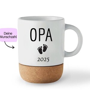 May include: White ceramic mug with a cork base. The mug features the word "OPA" in black, along with baby footprints and the year 2025. A pink label with German text is on the left side.