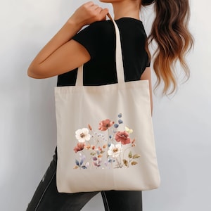 May include: Beige tote bag featuring a watercolor floral design with red, white, and blue flowers, and green leaves. The bag has long straps and is held over a person's shoulder. The bag is made of a canvas-like material.
