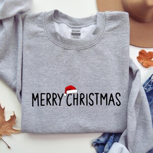 May include: A heather gray crewneck sweatshirt featuring the black text MERRY CHRISTMAS embroidered across the chest. A small red and white Santa hat is positioned directly above the letter I in the word CHRISTMAS.