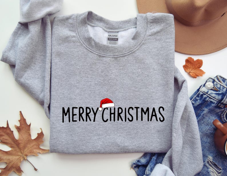 May include: A heather gray crewneck sweatshirt featuring the black text MERRY CHRISTMAS embroidered across the chest. A small red and white Santa hat is positioned directly above the letter I in the word CHRISTMAS.