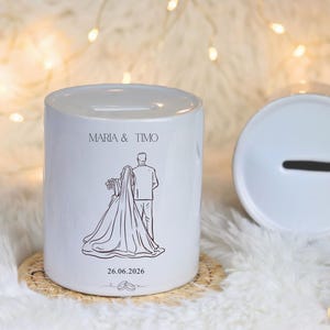 Puede incluir: Hucha de cer&aacute;mica blanca para bodas con una ranura en la parte superior. El banco presenta un dibujo lineal de una novia y un novio, los nombres "MARIA & TIMO" y la fecha "26.06.2026". El banco es cil&iacute;ndrico y se apoya en un posavasos tejido.
