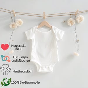 May include: A white baby onesie hangs from a wooden hanger, suspended by a clothes pin on a string. The onesie has short sleeves and snaps at the bottom. Text on the left side reads: "Hergestellt in DE", "F&uuml;r Jungen und M&auml;dchen", "Hautfreundlich", and "100% Bio-Baumwolle".