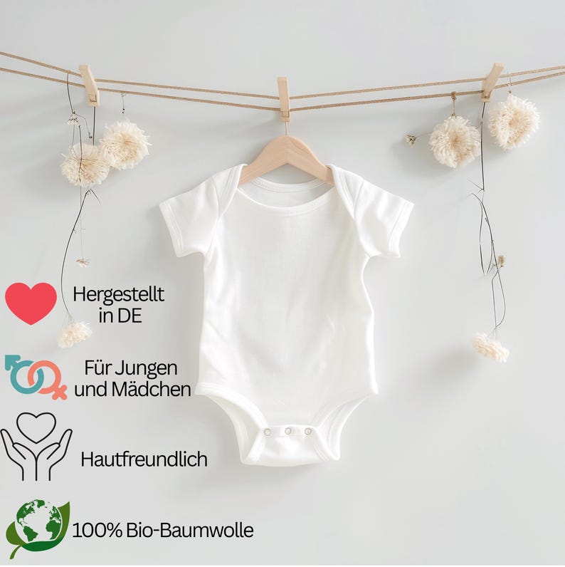 May include: A white baby onesie hangs from a wooden hanger, suspended by a clothes pin on a string. The onesie has short sleeves and snaps at the bottom. Text on the left side reads: "Hergestellt in DE", "F&uuml;r Jungen und M&auml;dchen", "Hautfreundlich", and "100% Bio-Baumwolle".