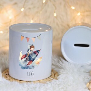 Op de afbeelding: Een witte keramische spaarpot met een cartoonillustratie van een astronaut op een raket. De spaarpot heeft een gleuf voor munten en de naam "LEO" op de voorkant gedrukt. Een tweede deksel is zichtbaar.