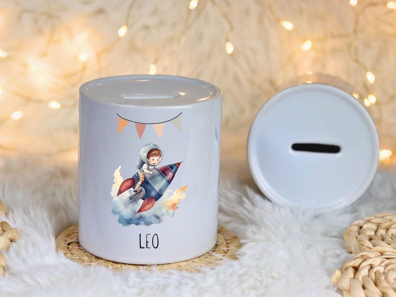Op de afbeelding: Een witte keramische spaarpot met een cartoonillustratie van een astronaut op een raket. De spaarpot heeft een gleuf voor munten en de naam "LEO" op de voorkant gedrukt. Een tweede deksel is zichtbaar.