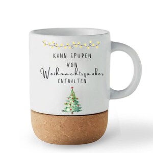 May include: White ceramic mug with a cork base, featuring a Christmas tree illustration and the text "Kann Spuren von Weihnachtszauber enthalten". The mug has a curved handle and a string of yellow lights.