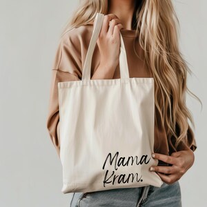 May include: Cream-colored tote bag with the words "Mama Kram" in black script. The bag has two long handles and is made of a sturdy fabric. The tote is held by a person with long blonde hair.