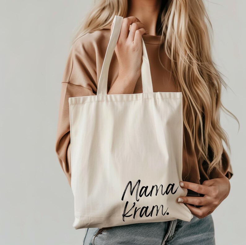 May include: Cream-colored tote bag with the words "Mama Kram" in black script. The bag has two long handles and is made of a sturdy fabric. The tote is held by a person with long blonde hair.