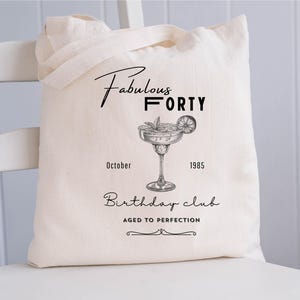 May include: A cream-coloured tote bag with black text and a cocktail illustration. The text reads "Fabulous Forty," "October 1985," and "Birthday club, Aged to Perfection." The bag has two handles and is on a white chair.