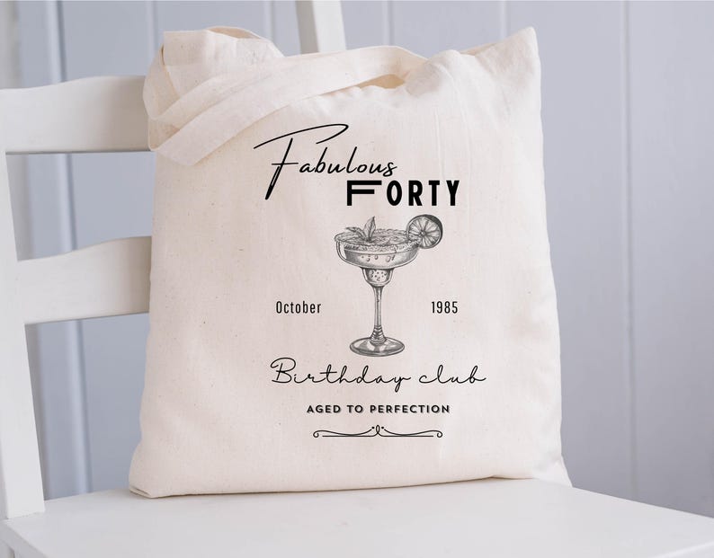 May include: A cream-coloured tote bag with black text and a cocktail illustration. The text reads "Fabulous Forty," "October 1985," and "Birthday club, Aged to Perfection." The bag has two handles and is on a white chair.