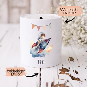 Op de afbeelding: Witte keramische spaarpot met een kleurrijke illustratie van een astronaut op een raket. De spaarpot heeft een gleuf aan de bovenkant en de naam "LEO" staat op de voorkant. Een banner met vlaggetjes staat boven de astronaut.