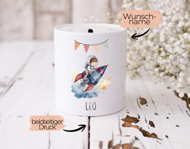 Op de afbeelding: Witte keramische spaarpot met een kleurrijke illustratie van een astronaut op een raket. De spaarpot heeft een gleuf aan de bovenkant en de naam "LEO" staat op de voorkant. Een banner met vlaggetjes staat boven de astronaut.