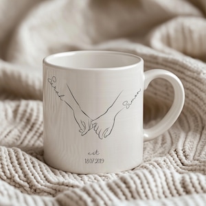 Könnte beinhalten: Weiße Keramiktasse mit einer schwarzen Strichzeichnung von zwei Händen, die sich am kleinen Finger halten. Die Namen "Sarah" und "Simon" sind auf der Tasse geschrieben, darunter das Datum "18.07.2019". Die Tasse hat einen gebogenen Henkel.
