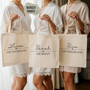 May include: Three beige tote bags with black script. Each bag has a name and title: "Luisa, DIE TRAUZEUGIN", "Sarah, DIE BRAUT", and "Sina, DIE TRAUZEUGIN". The bags are held by people wearing white robes.