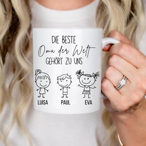May include: White ceramic mug with the text "DIE BESTE Oma der Welt GEH&Ouml;RT ZU UNS" and three stick figure illustrations with the names Luisa, Paul, and Eva. The mug has a white handle.