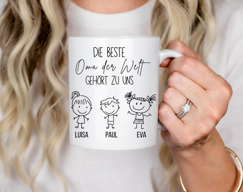May include: White ceramic mug with the text "DIE BESTE Oma der Welt GEH&Ouml;RT ZU UNS" and three stick figure illustrations with the names Luisa, Paul, and Eva. The mug has a white handle.
