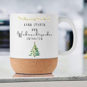 May include: White ceramic mug with a cork base. The mug features the text "Kann Spuren von Weihnachtszauber enthalten" and a Christmas tree illustration. A string of yellow lights is above the text. The mug is on a white surface.