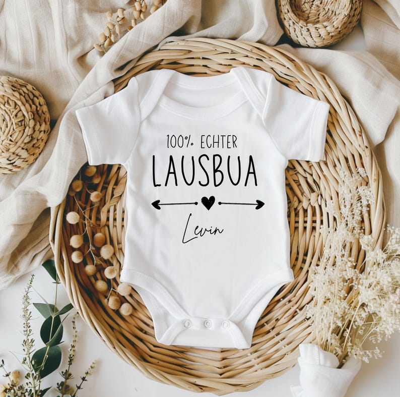 May include: White baby onesie with the text "100% ECHTER LAUSBUA" in black, with arrows and a heart. The name "Levin" is written below in a script font. The onesie is displayed in a woven basket.