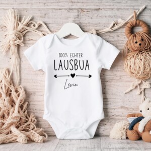 May include: White baby onesie with the text "100% Echter Lausbua" and the name "Levin" in black script. The design includes a heart and arrows. The onesie is hanging on a white hanger.