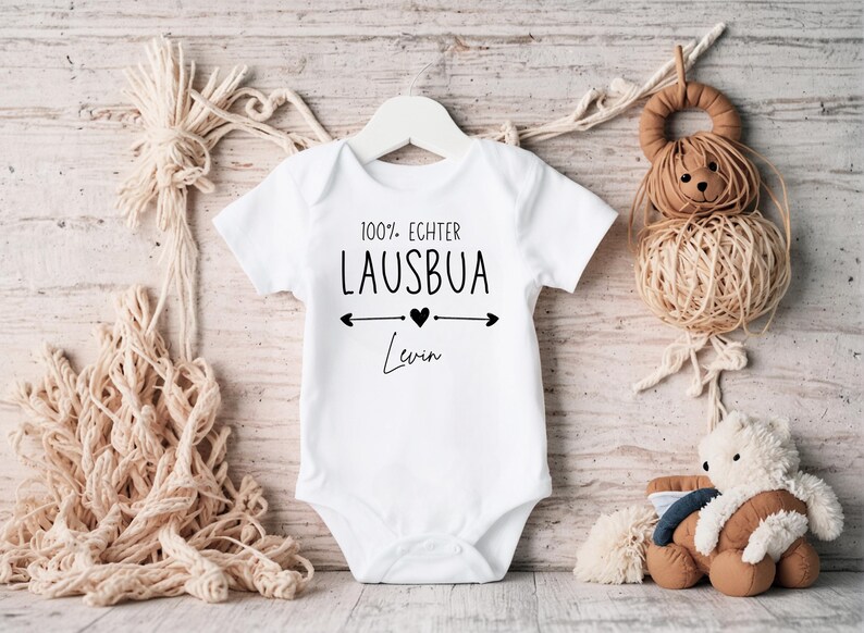 May include: White baby onesie with the text "100% Echter Lausbua" and the name "Levin" in black script. The design includes a heart and arrows. The onesie is hanging on a white hanger.