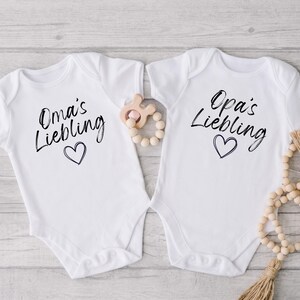 May include: Two white baby onesies with short sleeves. One onesie says "Oma's Liebling" and the other says "Opa's Liebling" in black script, each with a heart below. A wooden teething toy and a beaded garland are next to the onesies.