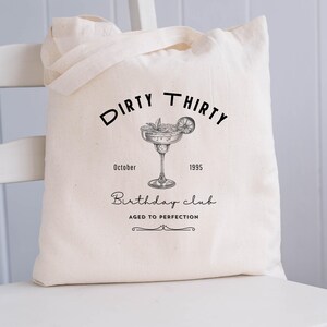 May include: Cream-colored tote bag with black text and a cocktail illustration. The text reads "DIRTY THIRTY Birthday club AGED TO PERFECTION" with the year 1995 and the month October. The bag has two handles.
