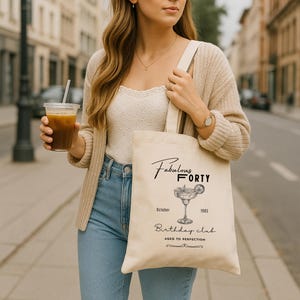 May include: A cream canvas tote bag with the text "Fabulous Forty" and "Birthday Club" printed in black. The bag features a margarita illustration and the dates "October 1985". The bag is carried by a person.