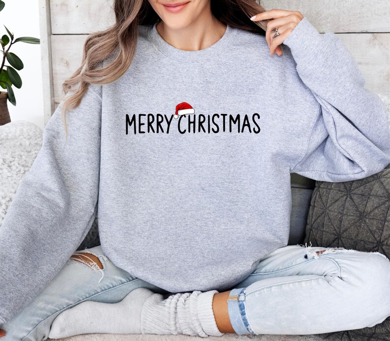 May include: A light gray crewneck sweatshirt featuring the black text MERRY CHRISTMAS printed across the chest. A small red and white Santa hat graphic is positioned directly above the letter I in the word CHRISTMAS.