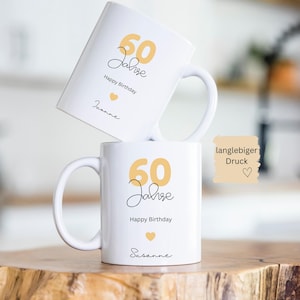 May include: Two white ceramic mugs with a birthday theme. The mugs feature the text "60 Jahre Happy Birthday" in black script and gold numbers. A small gold heart is also present. The name "Susanne" is written on one mug.
