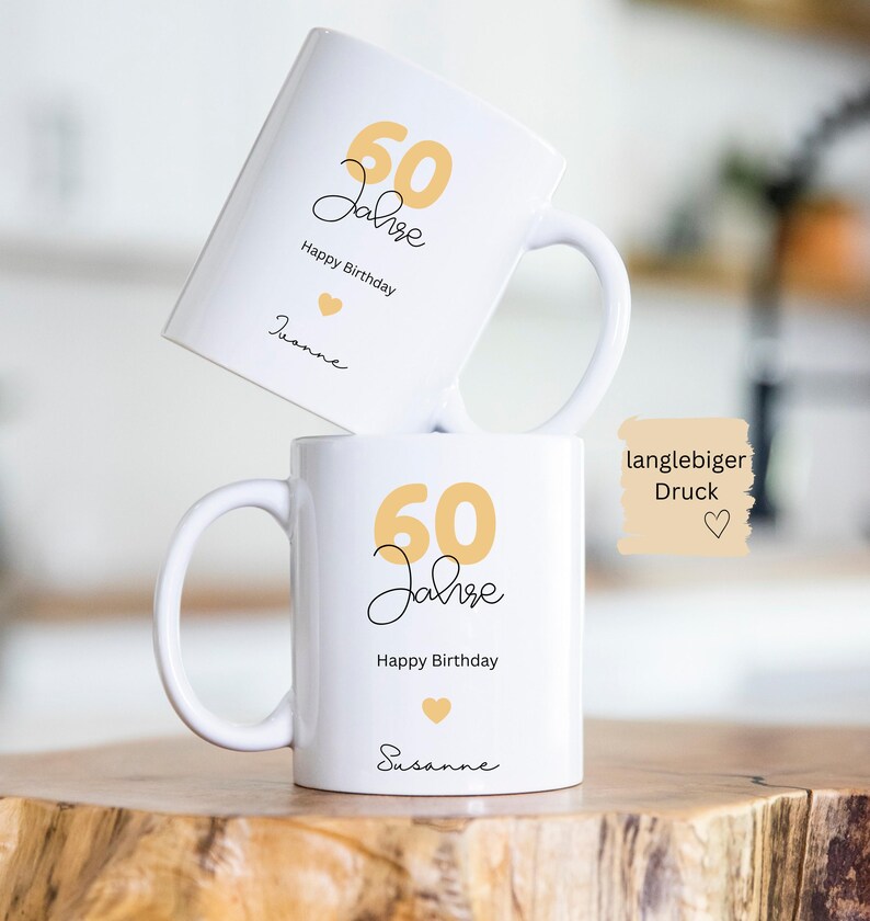 May include: Two white ceramic mugs with a birthday theme. The mugs feature the text "60 Jahre Happy Birthday" in black script and gold numbers. A small gold heart is also present. The name "Susanne" is written on one mug.