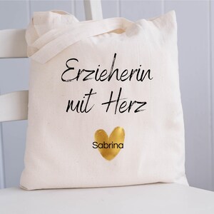 May include: A cream-colored tote bag with the text "Erzieherin mit Herz" in black script, a gold heart, and the name "Sabrina" below. The bag has two handles and is sitting on a white chair.