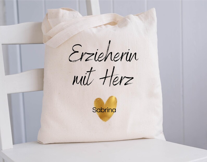 May include: A cream-colored tote bag with the text "Erzieherin mit Herz" in black script, a gold heart, and the name "Sabrina" below. The bag has two handles and is sitting on a white chair.
