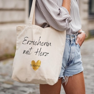 May include: Cream-colored tote bag with the text "Erzieherin mit Herz" in black script, a gold heart, and the name "Sabrina." The bag is carried over the shoulder.