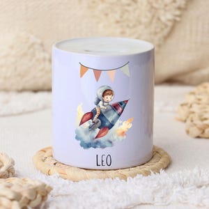 Op de afbeelding: Een witte keramische spaarpot met een cartoonillustratie van een astronaut op een raket. De raket heeft rode vlammen en is omgeven door wolken. De naam "LEO" staat eronder gedrukt. Bovenop staat een banner met oranje, roze en groene vlaggen.