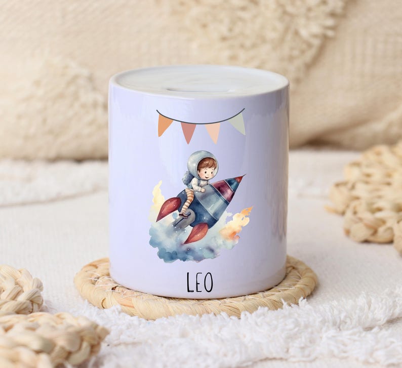 Op de afbeelding: Een witte keramische spaarpot met een cartoonillustratie van een astronaut op een raket. De raket heeft rode vlammen en is omgeven door wolken. De naam "LEO" staat eronder gedrukt. Bovenop staat een banner met oranje, roze en groene vlaggen.