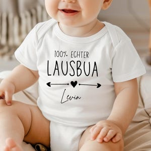 May include: A white baby onesie with the text "100% Echter Lausbua" in black, with a heart and arrows design. The name "Levin" is written below. The onesie is short-sleeved and is being worn by a baby.