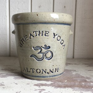 Pode incluir: Um pote de grés bege com as palavras "BREATHE YOGA" e o símbolo Om em azul. O pote tem duas pequenas alças e o texto "ALTON, NH" na parte inferior. O pote está sobre uma superfície de madeira branca.